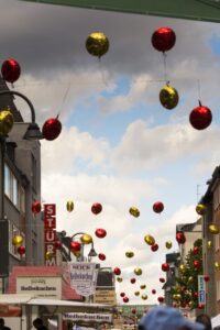 Straße mit Häusern rechts und links. Im bewölkten Himmel in der Mitte hängen an Drähten gelbe und rote Metallic-Luftballons.